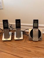 Three VTech cordless phone handsets placed on their respective base chargers on a wooden floor against a baseboard wall.