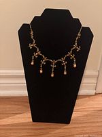 Full view of gold tone necklace with floral design and five dangling peach-colored pearls on necklace stand.