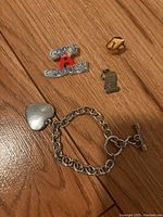 Photo of graduation enamel pin, gold tone pin, miniature scroll pin, and silver-tone chain heart charm bracelet on wood surface.