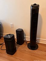 Photo of one tall black tower fan and two smaller black fans placed on wooden floor against wall with power outlet.
