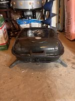 Black square charcoal BBQ grill with vent on lid and latch, viewed from the side showing legs and overall shape.