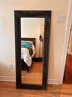 Large full body mirror with dark brown textured frame hung vertically showing reflection of bed and room.