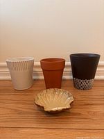 Three planting pots and one seashell-shaped dish arranged against a wall on a wooden floor.