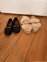 Black slip-on dress shoes and beige fluffy platform sandals side by side on wooden floor, front view