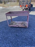 Steel utility cart with two levels, pinkish paint, and caster wheels visible outdoors on gravel surface.