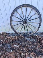 Two wagon wheels used for garden decoration, one visible propped against a white metal wall, with some dried leaves and gravel at the base.