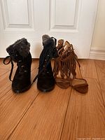 Front view of black winter boots with faux fur and tan suede heel sandals with fringe details on hardwood floor against white door