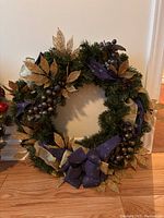 Christmas wreath with green pine foliage, gold glitter leaves, metallic berries, pine cones, and purple ribbon bow