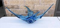 Front view of blue art glass stretch bowl with long pointed extensions on sides and center against brick wall background