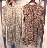 Front view of two Lost & Wander floral print long sleeve mini dresses hanging on wooden hangers, beige and dark floral prints visible