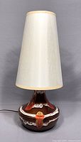 Full view of the lamp showing the cream-colored fabric shade and rounded pottery base with brown, white and orange glaze.