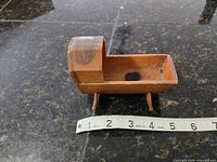 Miniature wooden cradle with measuring tape showing approximately 6 inch length on dark reflective surface.