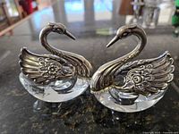 Front view of two swan-shaped salt cellars showing detailed silver wings and cut crystal bodies.