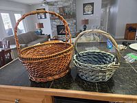 Two large woven baskets displayed side by side, one dark brown oval with twisted handle, the other grayish round shape with handle