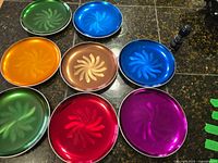 Nine multicolored round aluminum plates arranged on a dark surface showing swirling pattern design.