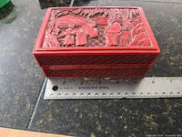 Front view of antique Chinese cinnabar box showing carved lid with figures and landscape, placed next to a steel ruler indicating width about 5.75 in.