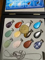 Open black display case showing 12 gemstone drop pendants arranged in foam with 18-inch silver-tone chain laid below them. A size reference coin and gemstone chart in lid.