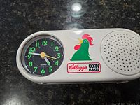 Front view of the Kellogg's advertising clock showing the round quartz clock face with green numbers and Kellogg's Corn Flakes rooster logo next to the speaker grill.