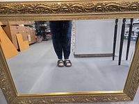 Front view of the large rectangular mirror with detailed gold frame reflecting floor and person wearing sandals.