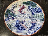 Top view of the large 12-inch round ceramic plate showing colorful koi fish swimming among water waves and a scenic natural background with trees and flowers.