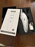 Opened box showing white Lucky Tail dog nail grinder in plastic wrap, user manual, and charger components in foam insert.