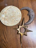 Three vintage garden art items on wooden floor: pottery moon plaque with dolphins, pottery crescent moon, solid brass star with circular center.