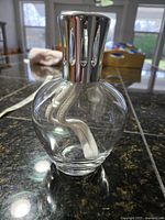Side view of the clear glass lamp body with white wick inside and attached silver-tone metal decorative top.