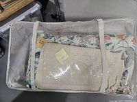 Two queen size comforters and one mattress protector stored in a transparent zippered bag. The comforters have floral patterns and the mattress protector is cream colored.