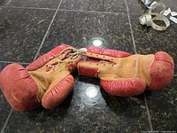 Photo of the pair of vintage child's boxing gloves, showing red and tan leather, laid on dark surface.