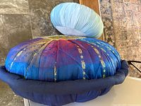 Photo showing both round yoga meditation pillows, a smaller light blue pillow and a larger ornate blue/purple patterned pillow with gold designs and navy trim.