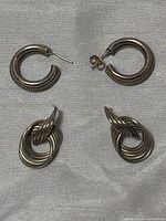 Two pairs of sterling silver stud earrings shown on fabric background. One pair is hoop style with ribbed texture, the other is a twisted intertwined loop design resembling a door knock.
