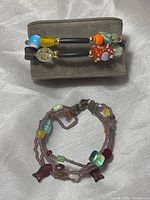 Two beaded bracelets on grey cushion: bottom one is multistrand fish net style with mixed beads including sterling clasp; top one is double band with colorful Murano glass and other beads.
