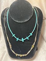 Two necklaces displayed on a black velvet bust. One necklace with turquoise beads and silver decorative beads. Second necklace with turquoise and off-white beads.