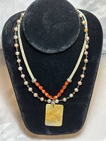 Two necklaces displayed on black necklace bust: one wired silver with alternating white and pinkish pearls; second silver leather cord with red coral beads and rectangular shell pendant