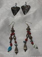 Two pairs of dangle earrings on gray cloth background. Top pair triangular oxidized sterling silver with small round opal stones. Bottom pair long dangles with mixed silver beads, red coral, turquoise beads, and white stone drops.