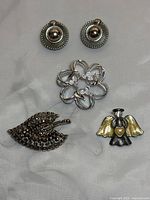 Four items: two round silver clip-on earrings, silver flower brooch, leaf brooch with stones, and enamel angel brooch with gold accents