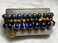 Three stretch gemstone bracelets stacked on a gray display cushion showing lapis lazuli, tiger's eye, and blue striped agate with CZ accents.