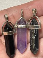 Three gemstone pendants shown held in fingers, colors visible as purple amethyst, solid black onyx, and black marble with white matrix, silver tone bails with partial cage setting.