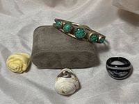 Lot of 3 rings and 1 bracelet arranged on light fabric background; rings include white plastic rose, black Murano glass with white and silver accents, and conch shell; bracelet is silver-plated with turquoise stones.