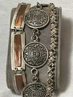 Three sterling silver bracelets displayed folded on a bracelet holder, showing a blend of metal textures and designs including Alpaca calendar medallions, copper panels, and a gemstone XO pattern bracelet.