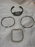 Four silver bracelets on beige fabric background. Two wavy plain bangles, one cuff with double oval design, one chain link bracelet.