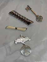 Photo showing all four silver brooch items arranged on a flat surface: skeleton key brooch, twisted bar brooch, white enamel bow brooch, Altar Society medal brooch