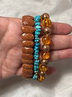 Hand holding three stretch beaded bracelets showing goldstone, turquoise chip, and copper/amber beads