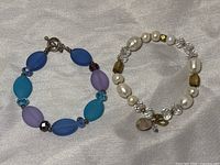 Two bracelets on a white cloth: One with multicolored glass oval beads, small faceted spacer beads, and toggle clasp. Second is a stretch bracelet with white pearls, clear crystal beads, olive green glass beads, and charms.