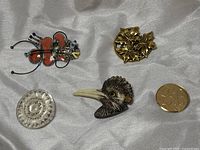 Five brooches arranged on a silk-like fabric: ceramic butterfly, floral gold metal round brooch, mother of pearl round brooch, carved resin bird head, and JB monogram metal leaf brooch with a coin for scale.
