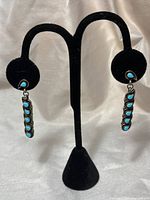 Pair of Native American sterling silver earrings displayed on black stand with visible turquoise inlays forming feather design, showing full length and shape.