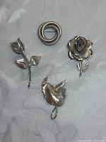 Five silver brooches arranged on fabric background including rose flower, circular spiral, leaves, and leaf designs.