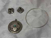 Photo of all three items: silver clip-on earrings, round pendant with turquoise and garnet stones, and plain silver bangle bracelet on a light fabric background.