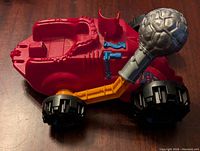 Photo showing the right side of the red He-Man Bashasaurus vehicle with visible details including an orange arm with a grey mace and black wheels.