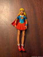 Front view of Supergirl figure wearing blue top with S emblem, red skirt and cape, and red sneakers posed standing on a wood surface.